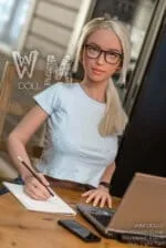 Sex Doll blond, Schulmädchen, schwarze Brille, grüne Augen, blaues geblümtes Oberteil, sitzt an einem Schreibtisch mit einem Bleistift in der Hand.