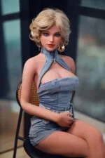 Sex Doll blondes kurzes Haar, blaue Augen, große Ohrringe, Jeanskleid, sitzt auf einem schwarzen Stuhl.