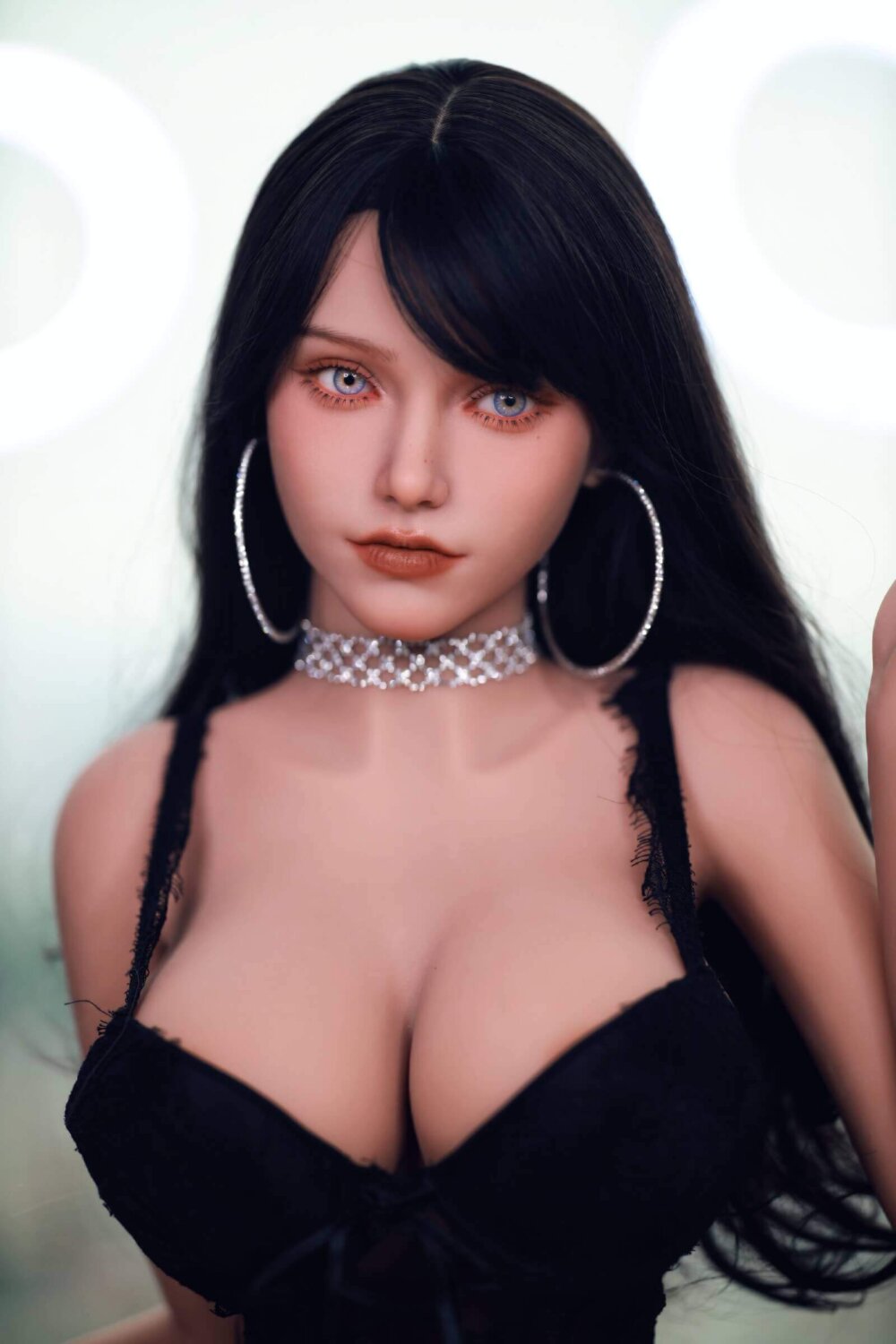 Sex Doll schwarzes Haar, große blaue Augen, silberne Halskette, große runde Ohrringe, schwarzes Oberteil.