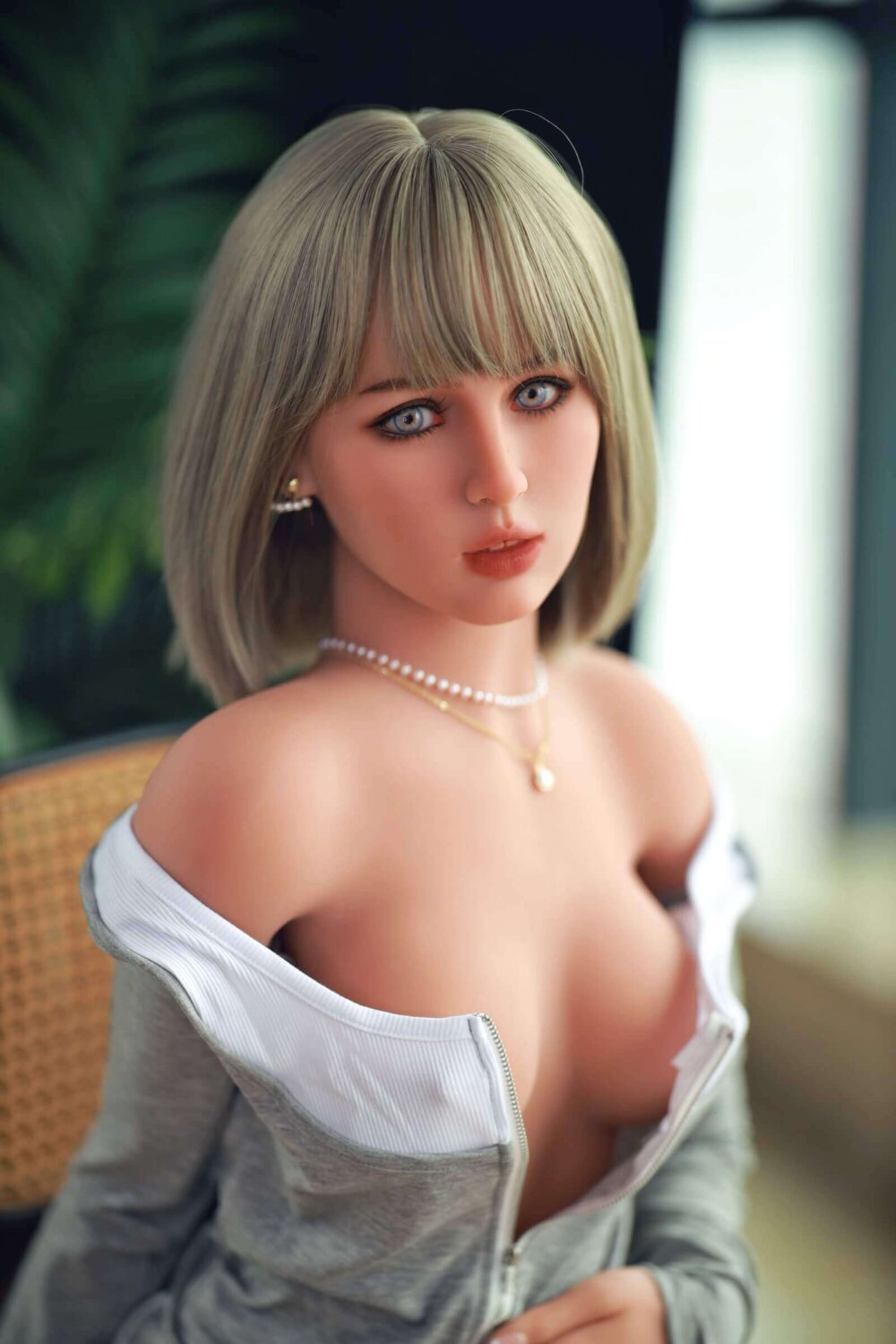 Sex Doll mit blonden Fransen, graues Sweatshirt mit Brüsten, Perlenkette und Ohrringe, blaue Augen.